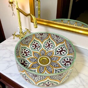 May include: A decorative ceramic vessel sink with a floral pattern in yellow, green, and burgundy. The sink has a gold-colored drain and is set against a white countertop. A gold faucet and mirror are visible in the background.