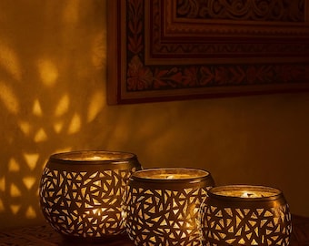 May include: Three golden candle holders with geometric cutouts, casting patterned light. The candle holders are placed on a wooden surface with intricate carvings. The warm lighting creates a cozy ambiance, perfect for home decor.