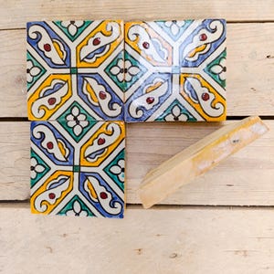 Puede incluir: Cuatro baldosas de cerámica decorativas con un colorido patrón geométrico en amarillo, azul, verde y rojo. Las baldosas son cuadradas y están dispuestas sobre una superficie de madera clara. También hay un trozo de madera rectangular.