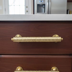 May include: Two ornate, gold-coloured cabinet pulls with a detailed floral pattern are mounted on dark brown wooden drawers. The pulls are cylindrical with decorative end caps and are attached to the drawer fronts with circular backplates.