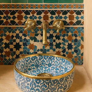 May include: A decorative bathroom sink with a blue and white floral pattern and a gold rim. The sink is mounted on a beige countertop, with a brass faucet and decorative tile backsplash in shades of blue, brown, and green.