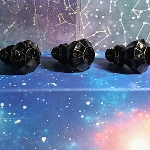 May include: Five faceted black glass bottle stoppers. The stoppers are arranged in a row on a blue surface with a starry sky design. The stoppers have a geometric design and a rounded top.