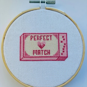 Könnte beinhalten: Ein rosa und weißer Kreuzstich-Stickrahmen mit dem Text "PERFECT MATCH" und einem rosa Herz in der Mitte.