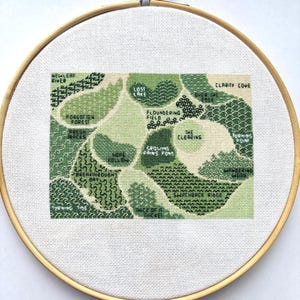 May include: A cross-stitch embroidery in a wooden hoop. The design features a map with various green shades, depicting locations like "Newleaf River," "Lost Lake," and "Clarity Cove." The text is stitched in white.