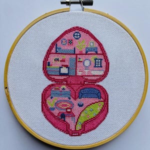 Puede incluir: Una bordado de punto de cruz rosa y azul de una casa de muñecas en miniatura con forma de corazón. La casa de muñecas tiene varias habitaciones, incluyendo un dormitorio, una cocina y un baño.