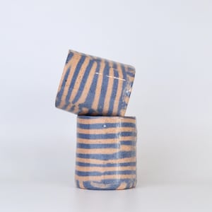 May include: Two cylindrical ceramic cups stacked vertically, featuring a speckled beige base with hand-painted horizontal blue stripes. The top cup is tilted at an angle, while the bottom cup sits upright, both displaying a smooth, glossy finish.