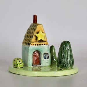 May include: A whimsical ceramic house figurine with a yellow star, a red door, and a green base. The house has a textured roof and is flanked by two green trees and a small, dotted green ball. The figurine is on a light green base.