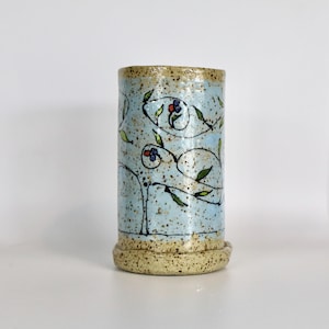 May include: A cylindrical ceramic vase with a light blue speckled background. The vase features a floral design with red and blue flowers, green leaves, and black stems. The rim and base are a speckled tan color.