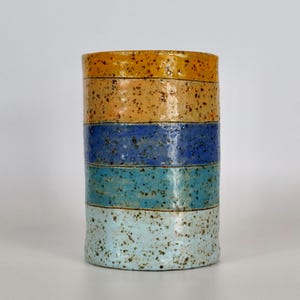 May include: A cylindrical ceramic vase with horizontal bands of color. The bands include shades of yellow, brown, blue, and turquoise, with a speckled texture. The vase is a decorative item.