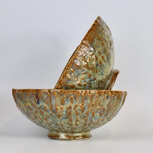 May include: Two ceramic bowls with a textured, uneven surface. The glaze features a mottled blend of metallic bronze, earthy brown, and muted sage green tones. One bowl sits upright on its base, while the second bowl rests tilted inside it.