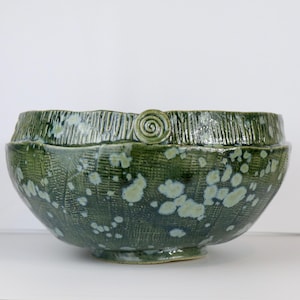 May include: A deep green ceramic bowl featuring a textured, cross-hatched surface pattern with light green mottled spots. The rim is accented with a raised, vertical ribbed texture and a single, prominent circular spiral detail on one side.