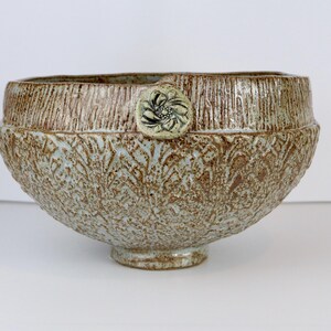 May include: A round ceramic bowl with a textured, mottled brown and light blue glaze. The exterior features a repeating embossed pattern and a smooth, circular medallion with a floral design attached to the upper rim of the vessel.
