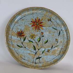 May include: A round ceramic plate with a hand-painted floral design. The plate features a light blue speckled background with orange, yellow, and green flowers. The rim is a tan color. The flowers are arranged in a circular pattern.