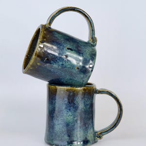May include: Two ceramic mugs stacked vertically, featuring a glossy glaze in shades of deep blue, teal, and earthy brown. The top mug is tilted at an angle, while the bottom mug sits upright. Both mugs have a curved handle and a smooth finish.
