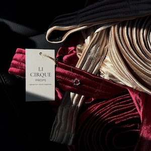 Puede incluir: Una colección de tiras de tela de terciopelo enrolladas en tonos burdeos intenso y oro metálico, con una etiqueta rectangular blanca sujeta con cordel. La etiqueta muestra el texto LI CIRQUE PROPS y texto adicional en caracteres cirílicos.