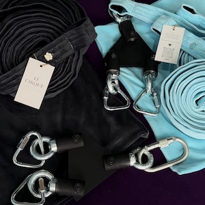 May include: Two aerial silks sets, one black and one light blue, each featuring a fabric length, a black triangular mounting plate, and silver metal carabiners. The black set includes a tag labeled LI CIRQUE, while the blue set has a small tag.