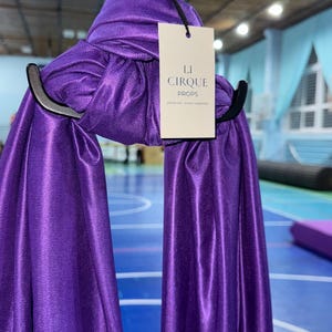 May include: A vibrant purple aerial silk fabric draped over a black metal rigging hardware piece. A rectangular white tag is attached to the fabric, featuring the black printed text LI CIRQUE PROPS. The fabric has a smooth, slightly reflective finish.