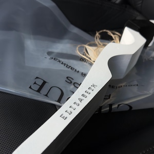 May include: A white, wooden bookmark with the name "ELIZABETH" stamped on it. The bookmark has a unique, curved design and a black accent at the top. It rests on a black leather surface.