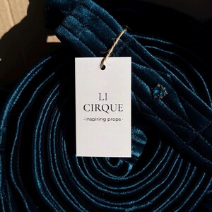 Peut inclure: Une longueur de tissu en velours bleu sarcelle profond enroulée, présentant une texture douce et pelucheuse. Une étiquette en papier blanc est attachée avec de la ficelle, portant le texte noir LI CIRQUE -inspiring props-. Un petit bijou bleu est visible.