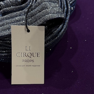 Puede incluir: Una tela gris oscuro enrollada con una textura brillante y brillante. Una etiqueta blanca adjunta dice "LI CIRQUE PROPS" con texto debajo. La tela está sobre una superficie púrpura.