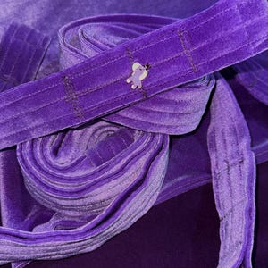 Peut inclure: Gros plan sur un tissu de velours violet foncé, avec une sangle assortie enroulée et une seconde sangle s'étendant du rouleau. Un petit charme en forme d'éléphant est cousu sur la sangle. Le tissu et les sangles sont d'un violet riche et uni.