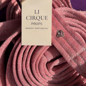 Puede incluir: Una longitud enrollada de tela rosa brillante con acabado metálico, que presenta un pequeño remache decorativo plateado. Se adjunta una etiqueta rectangular de color crema con el texto LI CIRQUE PROPS y una frase en cirílico.