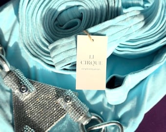 May include: A light blue aerial silk fabric folded into two circular coils, featuring a triangular metal rigging plate covered in silver rhinestones and two large silver carabiners. A white rectangular tag displays the text LI CIRQUE.