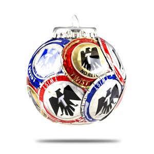 May include: A spherical ornament constructed from numerous metal bottle caps in red, white, blue, and gold. Each cap features a black or blue eagle emblem with a central letter T and the text TWIST or NO RETORNABLE. A small silver loop sits on top.