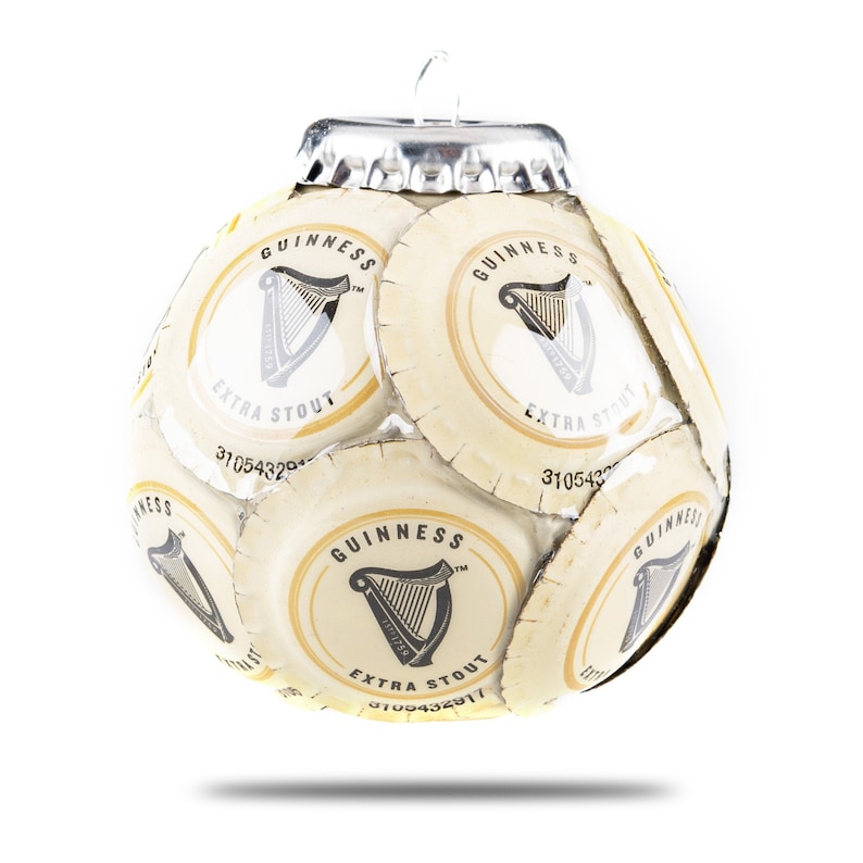 May include: A Christmas ornament shaped like a soccer ball made from Guinness beer bottle caps. The ornament is white with black and gold accents. The caps feature the Guinness logo, which includes a harp and the words "Guinness Extra Stout".