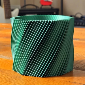 May include: A green, cylindrical planter with a unique, spiraling design. The planter is made of a smooth, textured material and has a wide opening at the top.