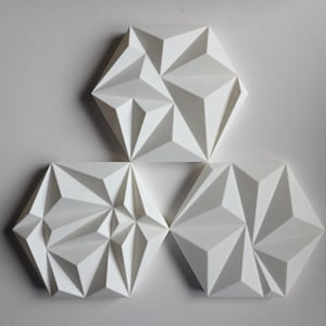 May include: Three white, geometric wall art pieces arranged in a triangular composition. Each piece is a hexagon with a complex, folded paper design, creating a play of light and shadow. The art is mounted on a plain white wall.