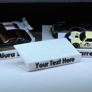 May include: A collection of toy cars, including a black Miura, a black and green car, and a yellow Carrera, displayed on white stands. A white sign in the foreground reads "Your Text Here" in black.