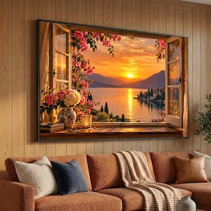 May include: A rectangular wall art print depicting an open window view of a sunset over a lake with mountains in the distance. The scene includes a vase of pink and white flowers, a small lit lantern, and a stack of books on the wooden windowsill.