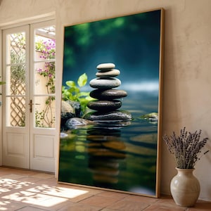 Könnte beinhalten: Ein großes rechteckiges Wandbild in einem dünnen goldenen Rahmen, das einen vertikalen Stapel aus glatten grauen und dunklen Steinen zeigt, die in flachem Wasser balanciert sind. Das Bild zeigt einen unscharfen grün-blauen Hintergrund mit Spiegelung.