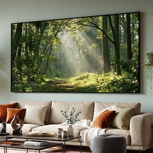 May include: A large rectangular wall art print featuring a sunlit forest scene with tall trees, lush green foliage, and light rays filtering through the canopy, displayed in a thin black frame above a beige sofa with various throw pillows.