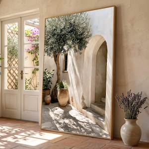 Op de afbeelding: Een grote rechthoekige vloerspiegel met een dunne gouden lijst. De spiegel reflecteert een mediterrane binnenplaats met een stenen boog, een olijfboom in een pot en een terracotta pot op een stenen terras met zonlicht en schaduwen.