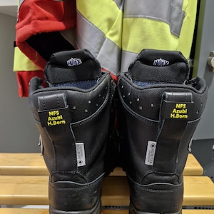 Op de afbeelding: Een paar zwarte leren brandweerlaarzen met gele en zwarte labels met de tekst "NFS Azubi H.Born" en "GORE-TEX CROSSTECH".