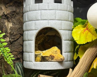 Op de afbeelding: Een lichtgrijs kasteelvormig reptielenverblijf met een gekuifde gekko erin. De gekko is geel en bruin. Het verblijf is omgeven door groene en gele bladeren en een digitale thermometer.