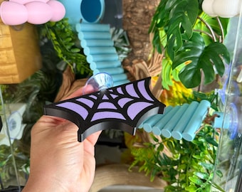 Op de afbeelding: Een zwart en paars spinnenwebvormige decoratie voor reptielenhabitats. De decoratie wordt voor een terrarium gehouden met een blauwe schuilplaats, een blauwe brug en kunstplanten. Het terrarium heeft een naturalistisch thema.
