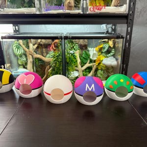 May include: Six colorful Pokeball-shaped containers with different designs. The containers are made of plastic and have a round shape with a small opening on the top. The designs include a black and yellow Pokeball, a pink Pokeball with a heart, a white Pokeball with a red stripe, a purple Pokeball with a white "M", a green Pokeball with yellow dots, and a red and blue Pokeball.
