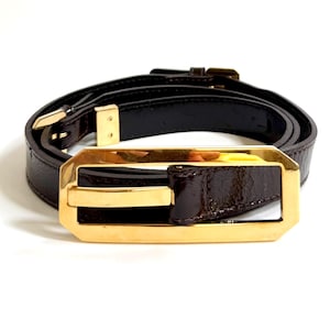 May include: A dark brown belt with a glossy finish, featuring a large, rectangular, gold-tone metal buckle with an open center design. The belt includes a matching dark brown leather-like strap, a gold-tone metal prong, and a small gold-tone keeper.
