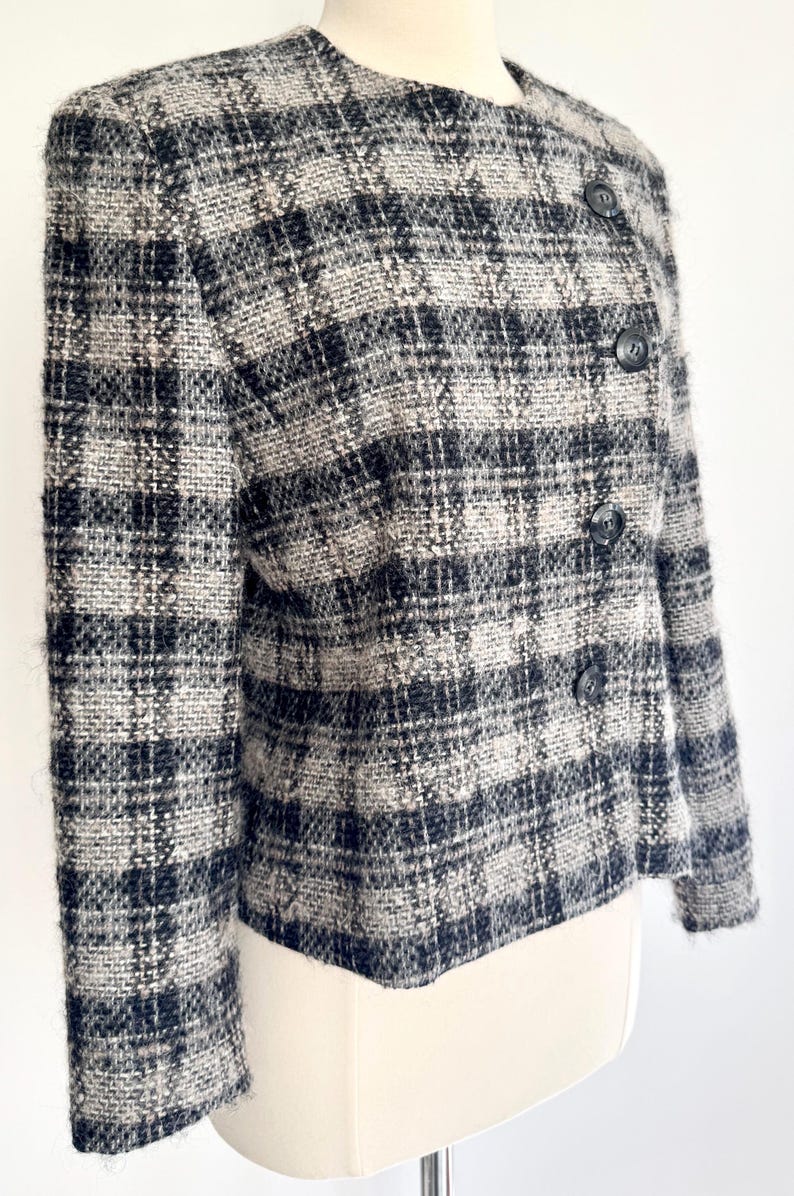 May include: A grey, black, and white checked jacket with a round neckline and three dark buttons. The jacket is made of a textured fabric and is displayed on a mannequin.