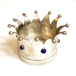 May include: A silver-toned crown-shaped vessel featuring a series of pointed arches along the top rim, each topped with a small spherical finial. The exterior band is decorated with several circular, deep blue cabochon stones set in metal.