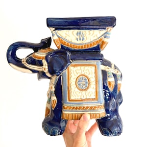 May include: A ceramic elephant garden stool in a vibrant blue hue, adorned with intricate white, tan, and gold details. The elephant features a rectangular top and decorative accents, showcasing a blend of artistry and functionality.