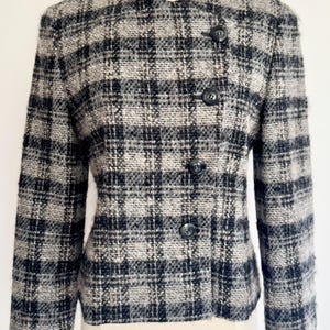 May include: A black, grey, and white checked jacket with a round collar and three black buttons. The jacket is made of a textured fabric and has long sleeves. The jacket is displayed on a mannequin.