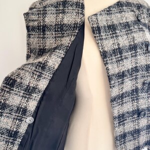 May include: A black, white, and beige plaid tweed jacket. The jacket features a round collar, three buttons, and a black lining. The garment is displayed on a mannequin.