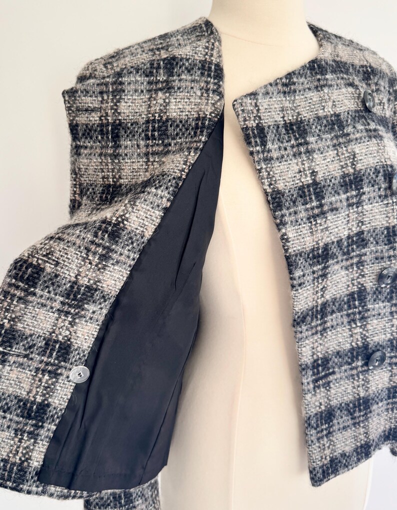 May include: A black, white, and beige plaid tweed jacket. The jacket features a round collar, three buttons, and a black lining. The garment is displayed on a mannequin.