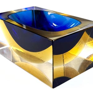 May include: A rectangular glass block bowl featuring a clear exterior and a deep blue interior basin. The thick glass construction creates a layered visual effect with a curved, semi-circular cutout design visible through the transparent sides.