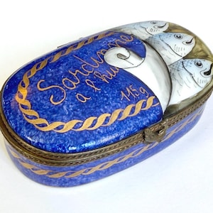 May include: An oval, blue and gold decorative box with the words "Sardines a l'huile" in gold script. The box features a design of sardines and a weight of 115g. The box is trimmed in gold and has a metal clasp.