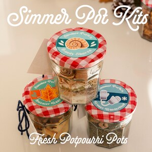 May include: Three glass jars filled with dried ingredients for simmer pot kits. Each jar has a red and white checkered lid with a circular label. Labels feature illustrations of a seashell, an orange maple leaf, and white mittens.